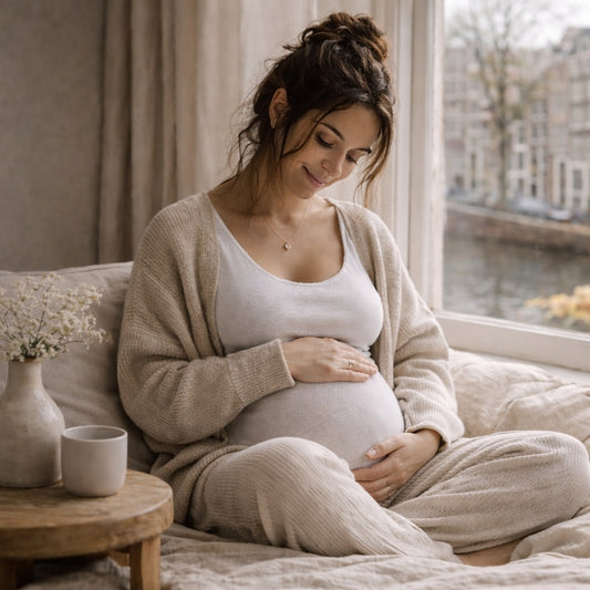 Zwangere vrouw in een beige vest zit bij het raam met haar handen op haar buik
